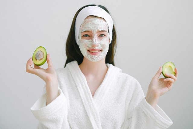 Lady wearing an avocado face mask in a white robe holding a halved avocado